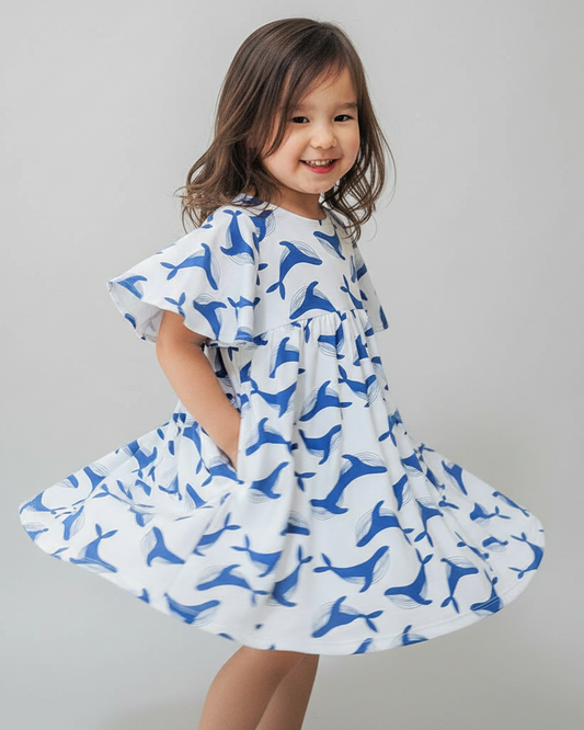  lively lifestyle scene featuring a young girl 3 year old with neat curls twirling in the 'We Fly High Dress - Blue Whale'  The camera angle is slightly elevated, capturing the dress's movement and the whimsical whale print vividly. Utilize an HDR 
