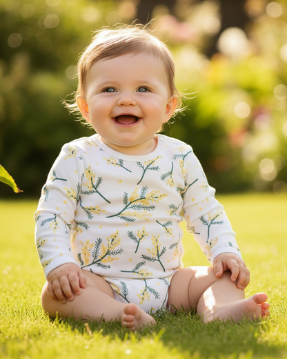 Baby sitting on grass wearing a wattle print onesie surrounded by greenery.