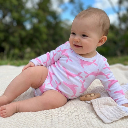 A lifestyle scene featuring a young baby wearing the Long Sleeve Bodysuit - Pink Whale with legs uncovered. The child is captured from a slight side angle, with a natural, candid expression. The background showcases softly blurred greenery and blue sky, wh